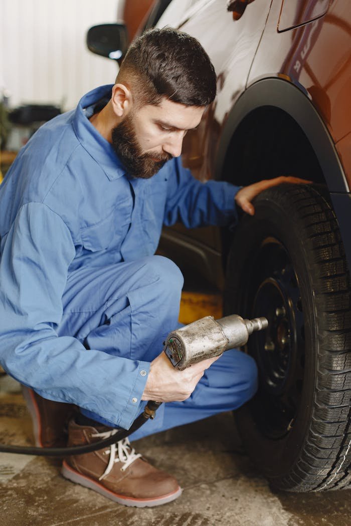 A mechanic in a blue uniform using a power tool to fix a car tire in a workshop indoors.